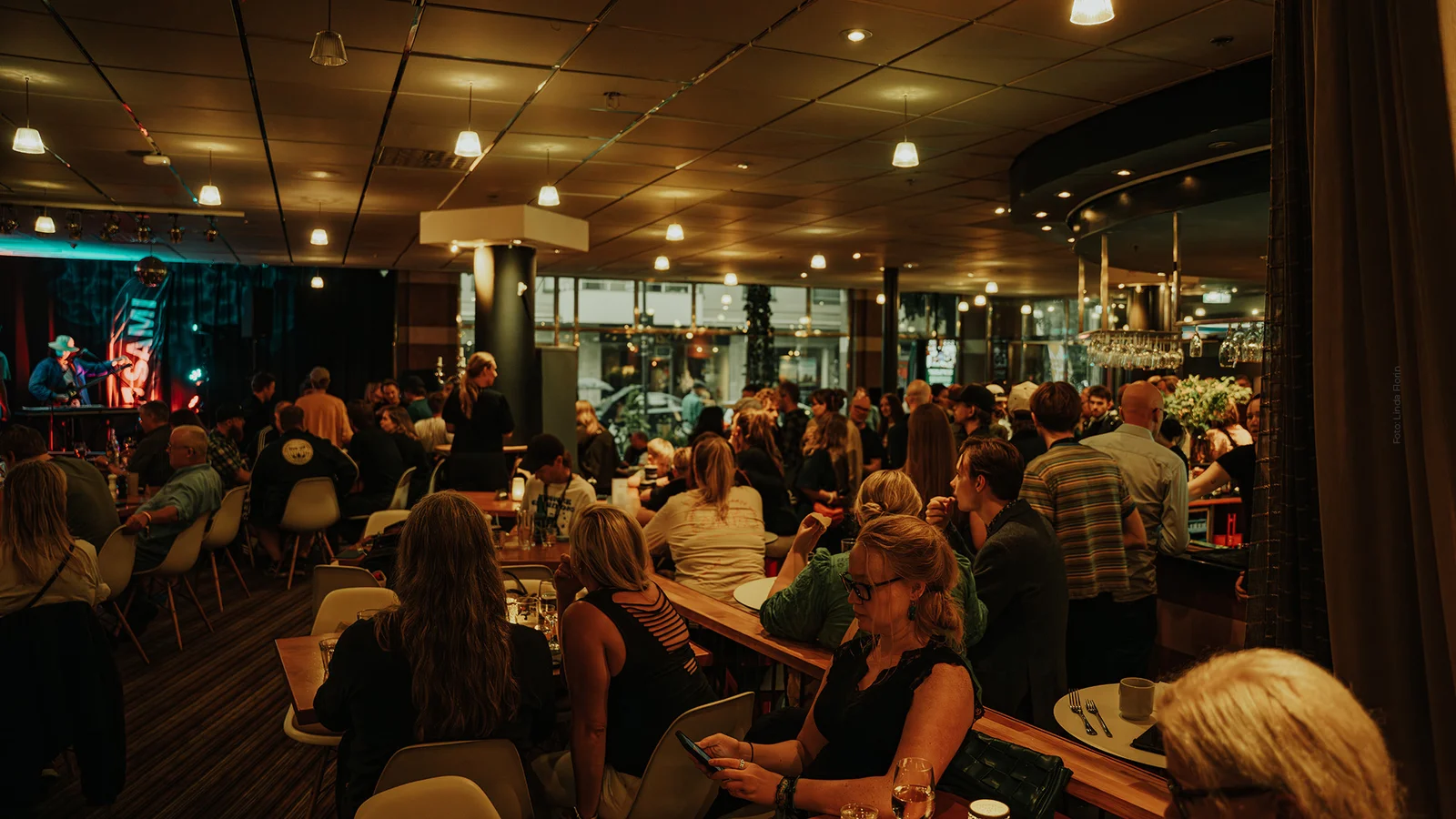 Stort öppen restauranglokal med scen till vänster och bar till höger fyllt av sittande gäster