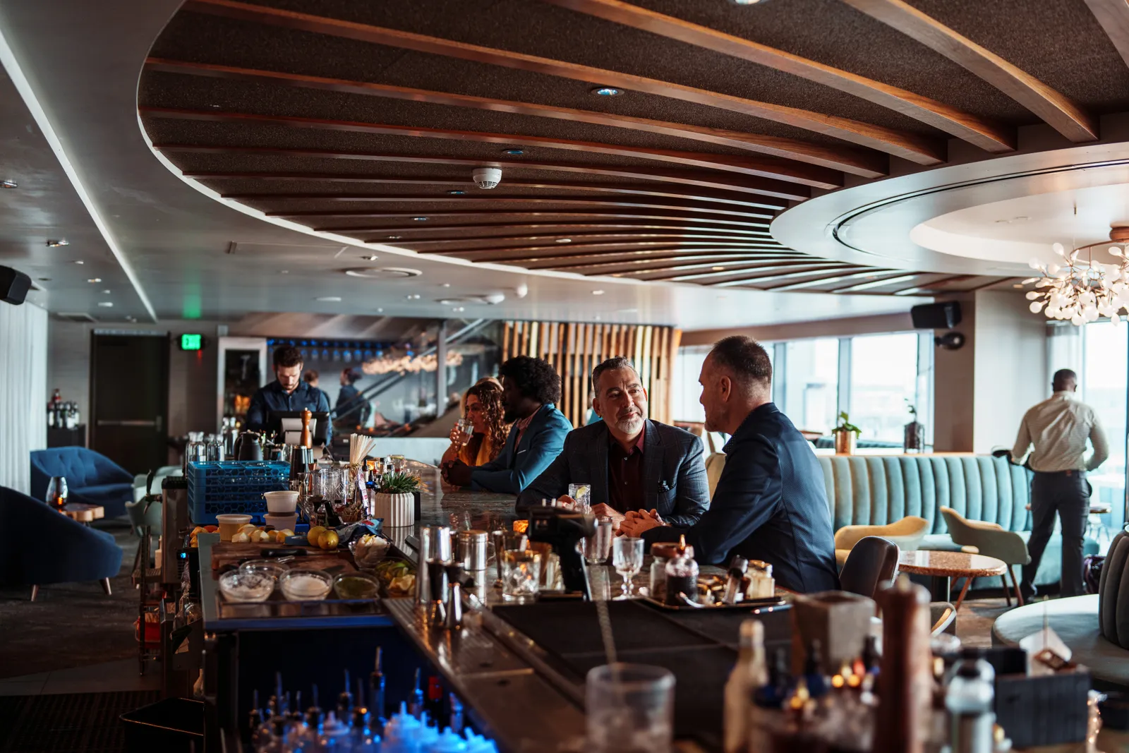 Personer sitter vid en modern bar med drinkar och bartendern förbereder beställningar i en stilfull lounge.