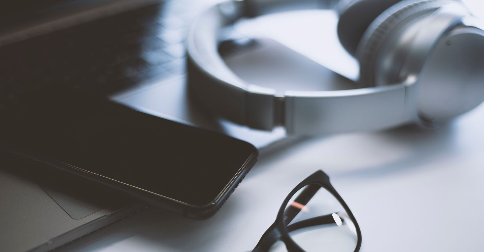 Närbild på telefon, glasögon, hörlurar och dator liggandes på ett bord
