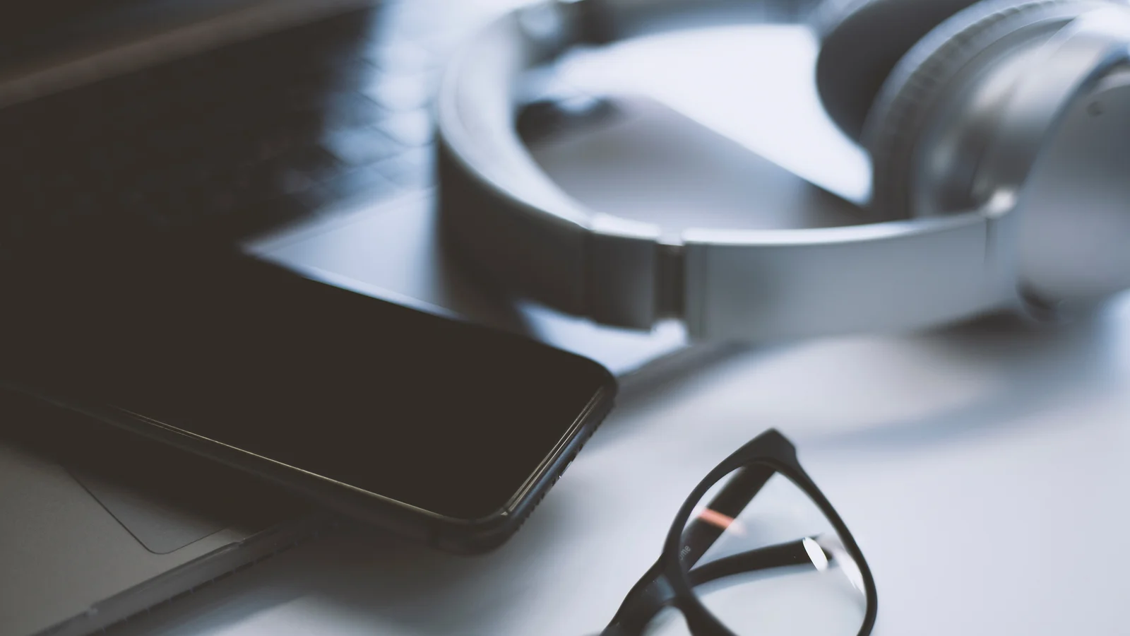 Närbild på telefon, glasögon, hörlurar och dator liggandes på ett bord