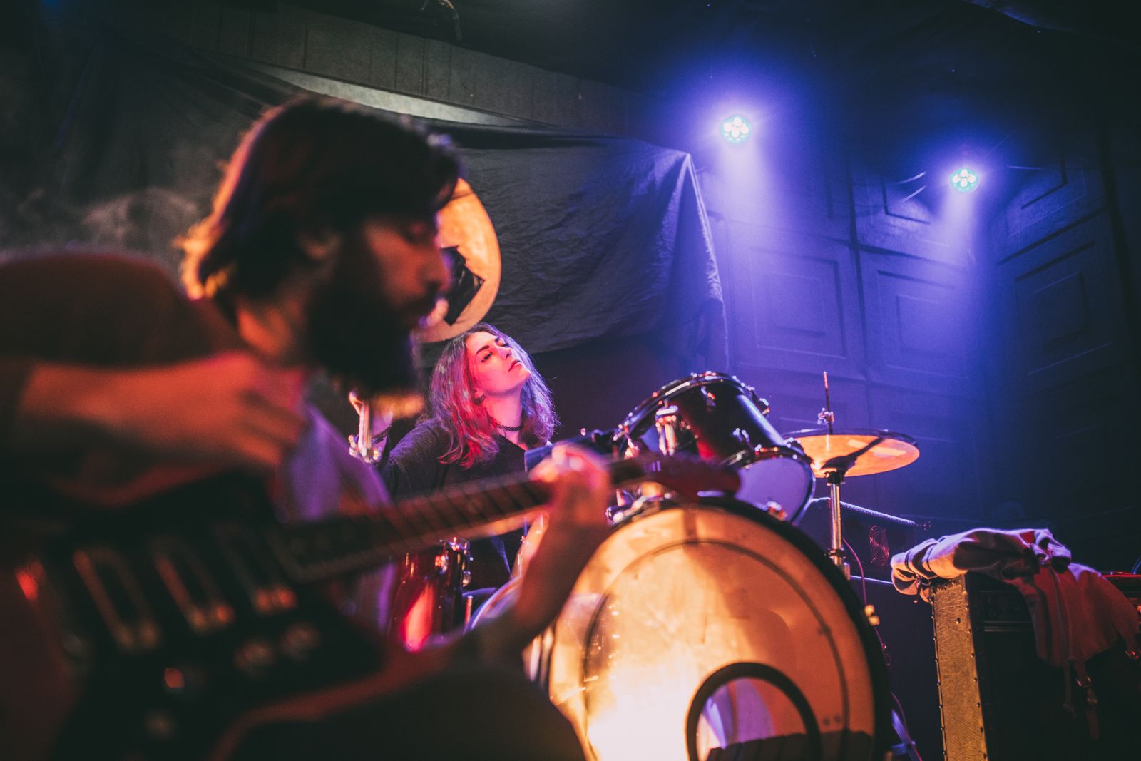 Gitarrist sittande i förgrunden och trummis i bakgrunden på scen med scenbelysning i lila