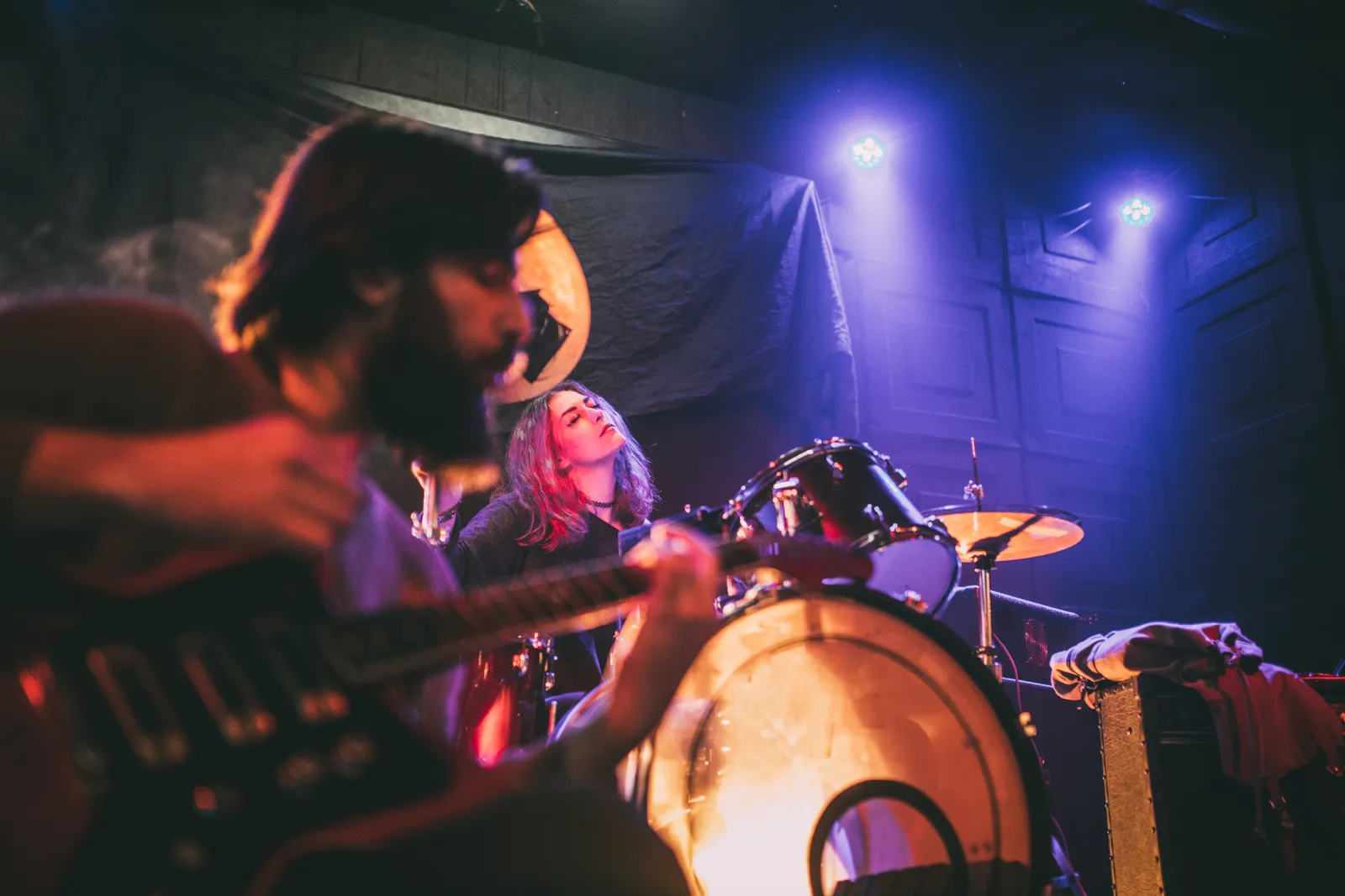 Gitarrist sittande i förgrunden och trummis i bakgrunden på scen med scenbelysning i lila