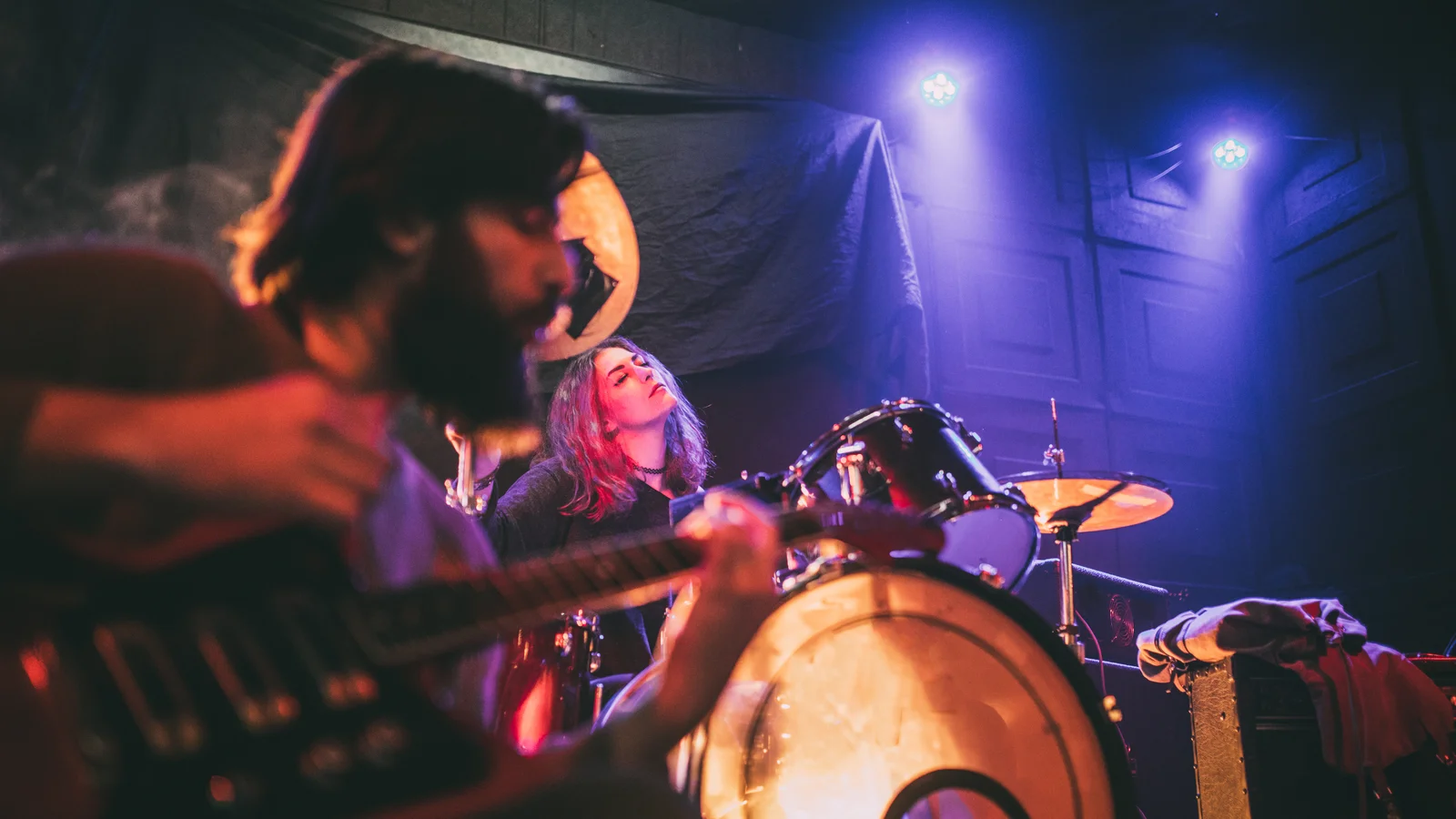 Gitarrist sittande i förgrunden och trummis i bakgrunden på scen med scenbelysning i lila