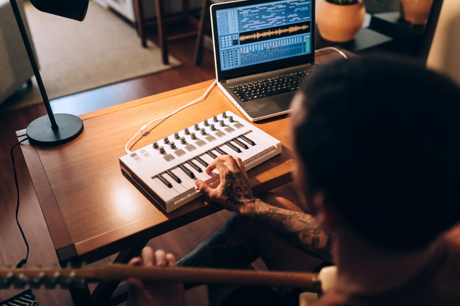 Person som sitter med gitarr i förgrunden med en hand på en mini keyboard och lampa samt dator med musikproduktionsmjukvara i bakgrunden