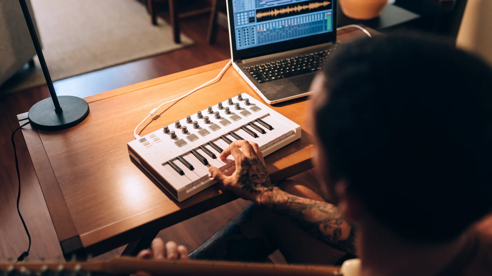 Person som sitter med gitarr i förgrunden med en hand på en mini keyboard och lampa samt dator med musikproduktionsmjukvara i bakgrunden